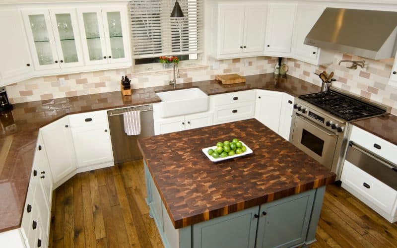 A clean fully equipped kitchen surrounded with cabinets and a square island in the middle with a tray of green fruit in the middle.